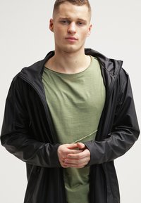 Musta, kevyt, vedenpitävä takki, jossa on huppu. Pitkät hihat elastisilla resoreilla, kerrostettuna vihreän, pyöreäkauluksisen T-paidan päälle.