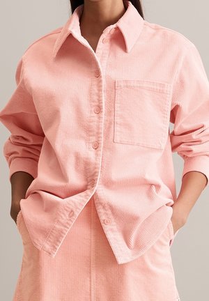 Person iført en lyserød fløjlsbutikskjorte med brystlomme, ærmerne skubbet op, og matchende lyserøde fløjlsbukser med hænderne i lommerne.