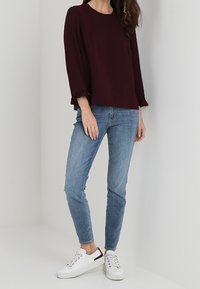 Eine Frau trägt ein dunkelburgunderfarbenes Langarmoberteil mit Rüschensaum, helle blaue Slim-Jeans und weiße Sneakers mit schwarzen und roten Akzenten.