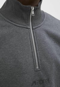 Gros plan sur un pull gris foncé à fermeture éclair quart de longueur, avec col côtelé et fermeture éclair métallique fermée, montrant une partie de la zone de la poitrine.