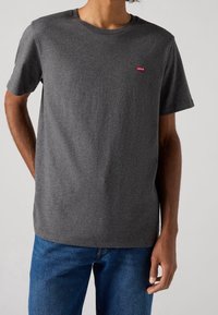 T-shirt en coton gris à manches courtes et col rond. Présente un petit logo rouge Levi's sur la poitrine gauche. Porté avec un jean bleu.