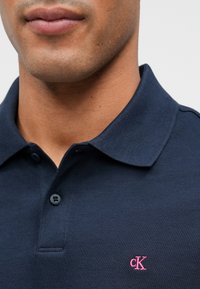 Námořní modré polo tričko z texturované látky, s límcem na dva knoflíky a růžovým logem "cK" na levém prsu.