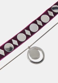 Silberner geometrischer Anhänger an einer metallischen Kette, der ein kreisförmiges Design zeigt und mit einem lila Samtband verziert ist, das glänzende kreisförmige und diamantförmige Elemente aufweist.