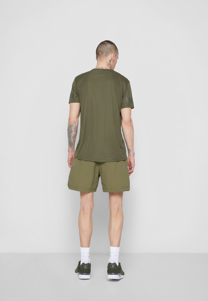 Oliwkowy t-shirt sportowy z krótkimi rękawami, w połączeniu z pasującymi szortami. Teksturowana tkanina, prosty krój oraz białe skarpetki z zielonymi butami.