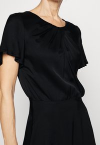Černé saténové šaty s krátkými volánkovými rukávy, kulatým výstřihem, plisovanými detaily na hrudi a přiléhavým pasem.