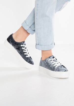 Persoon die opgerolde lichtblauwe spijkerbroek en grijze veterplateau sneakers met fluweelachtige textuur draagt, staand op een witte vloer.