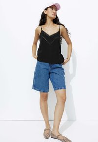 Camisole noire avec dentelle, shorts en denim bleu et ballerines à imprimé léopard. Modèle portant une casquette rose, debout devant un fond blanc.