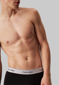 Fekete pamut boxer alsónadrág szürke derékrésszel, amelyen a "Calvin Klein" felirat szerepel. A törzsön bőr látható, semleges háttérrel.