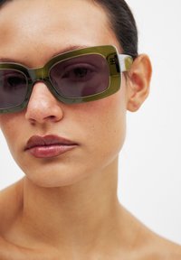 Zaļas pārmērīgi lielas saulesbrilles ar taisnstūra rāmjiem un tumšiem stikliem, ar caurspīdīgu akcentu ap malām un gludu apdari.