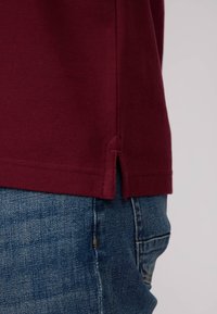 Polo en bordeaux avec un tissu texturé, doté de fentes sur l'ourlet inférieur, associé à un jean en denim bleu.
