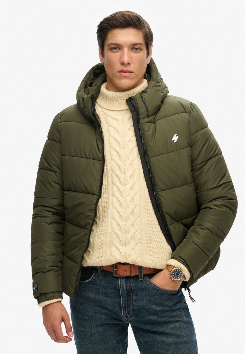 Olivegrøn pufferjakke med lynlåslukning, fast hætte og sorte detaljer; båret over en cremefarvet kabelstriktrøje og blå jeans.