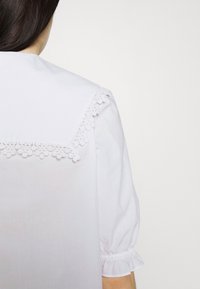 Femme portant un chemisier blanc avec un empiècement en dentelle à l'arrière et des manches courtes bouffantes à poignets élastiques, vue de dos.