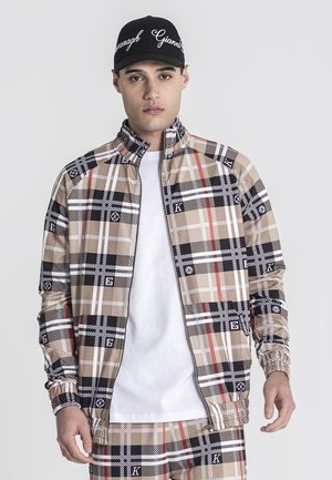 Chaqueta de cremallera a cuadros beige y negra con acentos rojos y puños elásticos, combinada con una camiseta blanca sencilla y una gorra negra con texto blanco.
