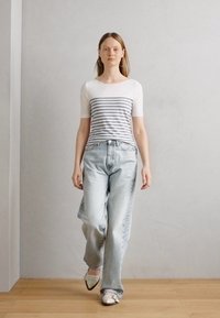 Pruhovaný top s krátkym rukávom v bielej a šedej farbe, skombinovaný s voľnými svetlo modrými džínsami. Model má na sebe biele topánky; pozadie je neutrálne.