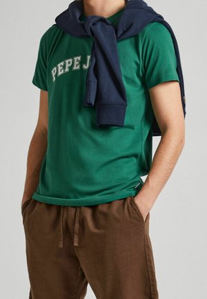 Vihreä puuvillapaita, jossa on valkoisella painettuna "PEPE JEANS". Laivastonsininen collegepaita sidottuna kaulalle. Ruskeat rento-mitoituksessa olevat housut taskuilla.