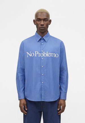 Homme aux cheveux blonds courts portant une chemise bleue à rayures blanches avec l'inscription « No Problemo » sur la poitrine, debout devant un fond blanc.