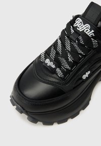 Μαύρο αθλητικό παπούτσι Buffalo με χοντρή σόλα, μαύρα και άσπρα στοκάρτα κορδόνια και ορατά λογότυπα της μάρκας στη γλώσσα, στην ετικέτα των κορδονιών και στο πλάι.