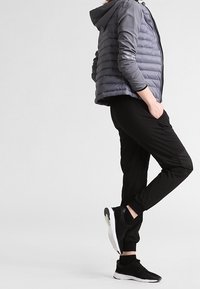 Doudoune grise avec capuche, pantalon de jogging noir avec des détails perforés, et chaussures de sport noires avec semelles blanches.