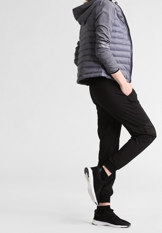 Doudoune grise avec capuche, pantalon de jogging noir avec des détails perforés, et chaussures de sport noires avec semelles blanches.