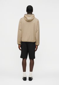 Άνδρας που στέκεται με την πλάτη γυρισμένη φορώντας μπεζ μπουφάν με κουκούλα, μαύρα σορτς μέχρι το γόνατο, λευκές κάλτσες και μαύρα παπούτσια μπροστά σε απλό φόντο.