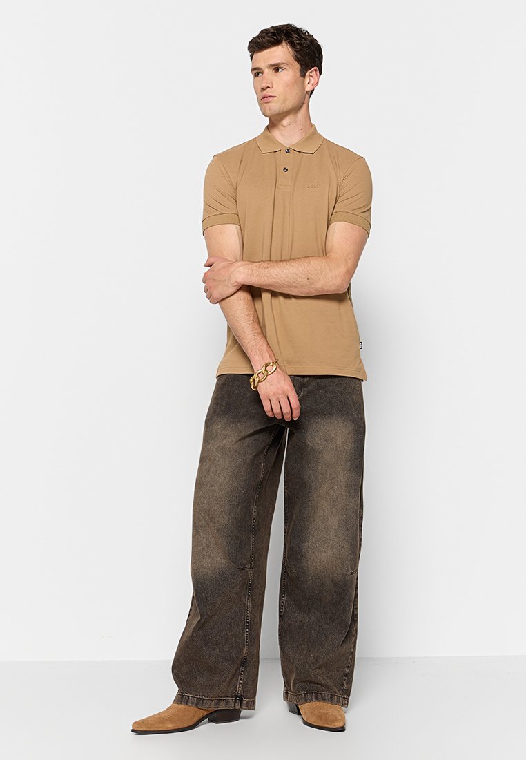 Polo shirt tan à manches courtes, avec un placket à deux boutons, associé à un jean en denim sombre, ample, et des bottes marron clair.