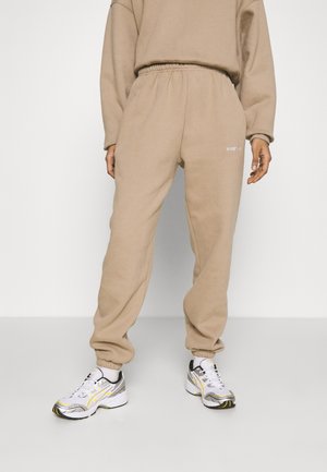 Personne portant un pantalon de survêtement beige et un sweat assorti avec des baskets blanches et argentées, debout sur un fond blanc.
