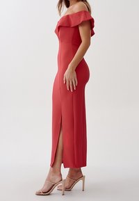 Rød off-the-shoulder kjole med flæseomgang, tætsiddende silhuet, sideslids og tekstureret stof. Båret med nude høje sandaler.