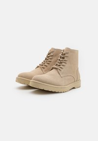 Botas de tobillo de ante beige con punta redonda, parte delantera con cordones y upper texturizado. Presenta una suela de goma gruesa con un patrón de relieve.