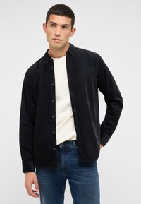 Schwarzes Cordhemd mit Knopfverschluss, langen Ärmeln und klassischem Kragen, über einem cremefarbenen T-Shirt getragen und mit blauen Jeans kombiniert.