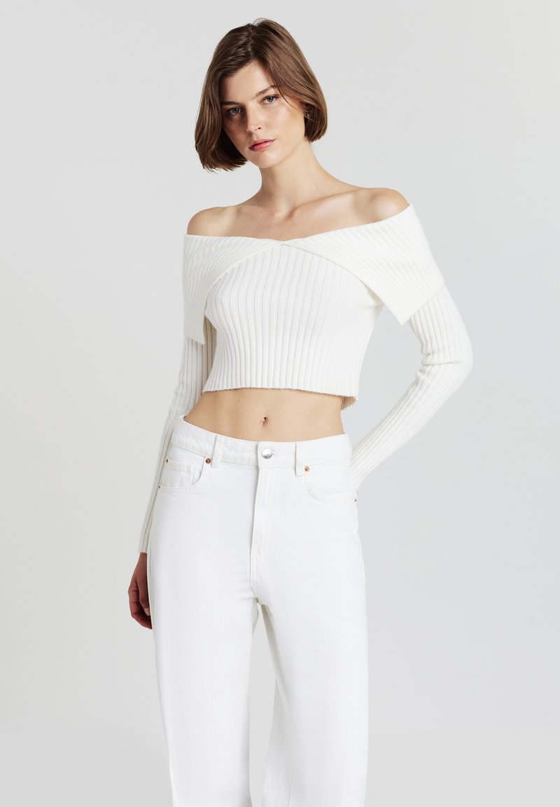 Weißer, schulterfreier, gerippter Crop-Pullover mit langen Ärmeln und einem überkreuzten Design. Kombiniert mit weißen, hochgeschnittenen Jeans.