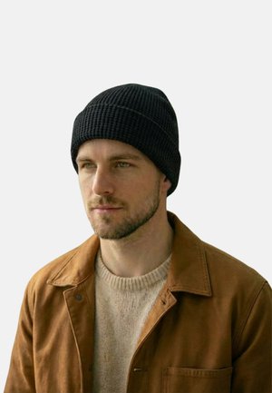 Mann mit schwarzer Strickmütze, beigem Pullover und brauner Jacke, der leicht nach links blickt vor einem schlichten hellen Hintergrund.