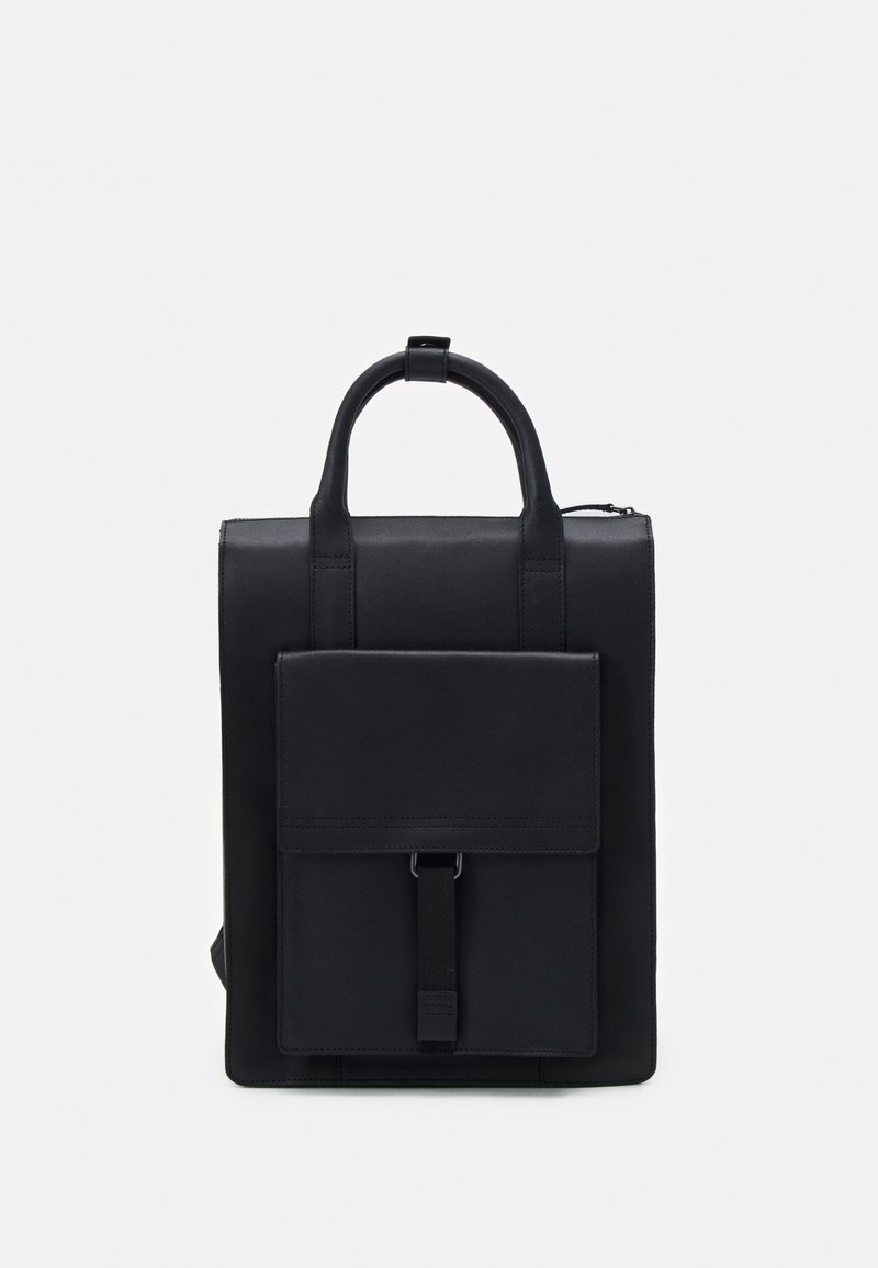 Sac à dos en cuir noir de forme rectangulaire, avec une poignée supérieure et une poche avant à rabat maintenue par une sangle. Texture lisse, design minimaliste.