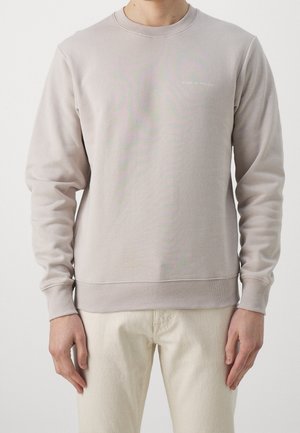 Mies pukeutuneena vaaleanbeigeen, pitkähihainen, pyöreäkauluksinen pusero, jossa pieni "Tiger of Sweden" -logo rinnassa, ja kermanväriset housut.