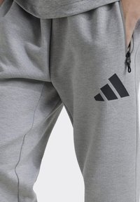 Szürke jogger nadrág, puha anyagból készült, cipzáros zsebbel és fekete Adidas logóval, laza szabású és oldalsó varrással.