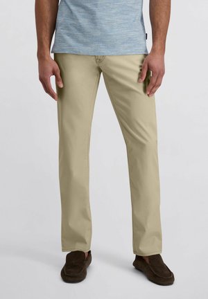 Homme portant un pantalon beige droit, une chemise bleu clair chinée et des mocassins marron, debout devant un fond uni.