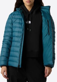 Una persona che indossa una giacca trapuntata color teal aperta su un hoodie nero con un logo bianco, pantaloni neri, in piedi contro uno sfondo bianco.