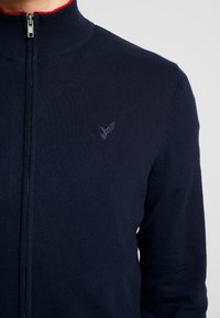 Marineblå zip-up tröja med hög krage, ribbad struktur och broderad fåglogotyp. Har en kontrasterande röd innerkrage.