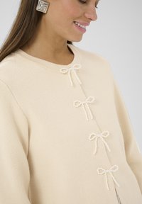 Beige kötött pulóver, amely gyöngyös masnikkal díszített elülső részen, kerek nyakkivágással és laza szabással, finom textúrával.