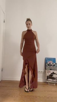Abito maxi ruggine con scollatura a halter, corpetto aderente e orlo asimmetrico con volant. Indossato con infradito nere. Sfondo semplice.