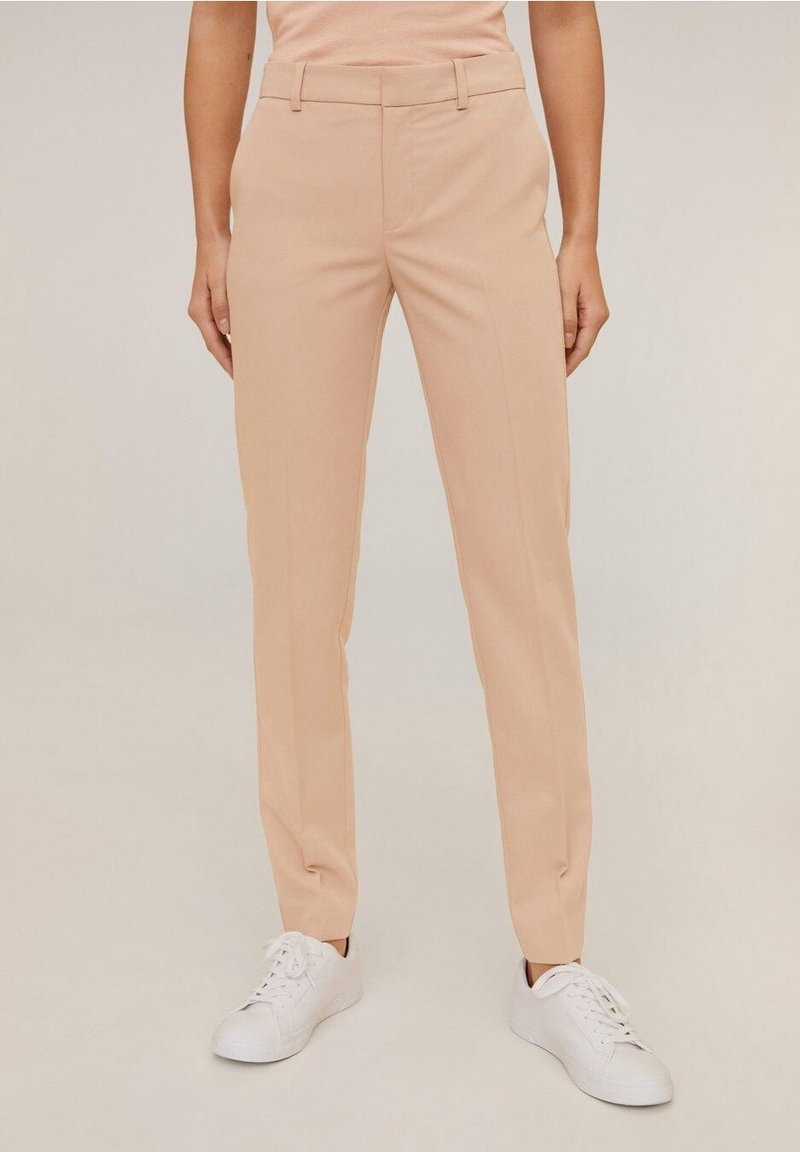 Femme portant un pantalon droit beige et des baskets blanches, debout devant un fond clair uni.