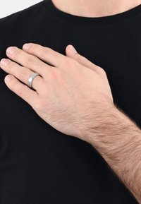 Un primer plano de una mano con un anillo de plata descansando sobre una camiseta negra, mostrando un gesto de la mano sobre el área del pecho.