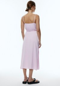Helles rosa, ärmelloses Midikleid mit geradem Schnitt, verstellbaren Trägern und seitlichem Schlitz. Das Model kombiniert es mit schwarzen Plateausandalen.