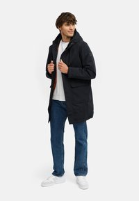 Zwarte gewatteerde parka-jack met twee voorkonten, verstelbare trekkoorden en een ritsluiting. Draagt over een witte shirt en denim jeans.