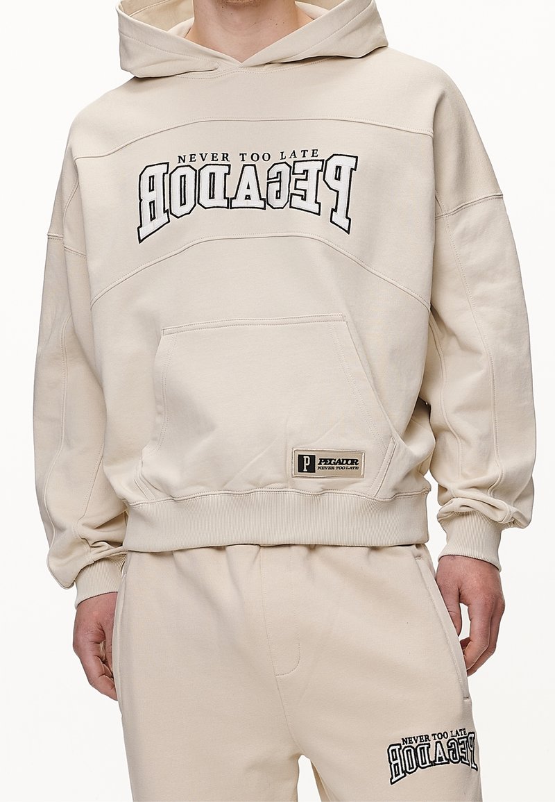 Homme portant un sweat à capuche beige et un pantalon de survêtement assorti avec le texte "PEGADOR NEVER TOO LATE" en orientation inversée et normale.