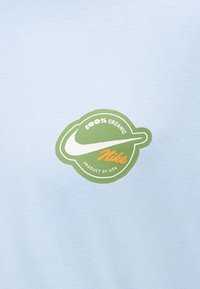 Λογότυπο Nike με κείμενο "100% Οργανικό" και "Προϊόν των ΗΠΑ" μέσα σε πράσινο κυκλικό σήμα σε φόντο από ανοιχτό γαλάζιο ύφασμα.