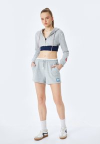 Młoda kobieta stojąca z rękami w kieszeniach, ubrana w szary, krótki hoodie, granatowy stanik sportowy, szare szorty, białe skarpetki i białe sneakersy.