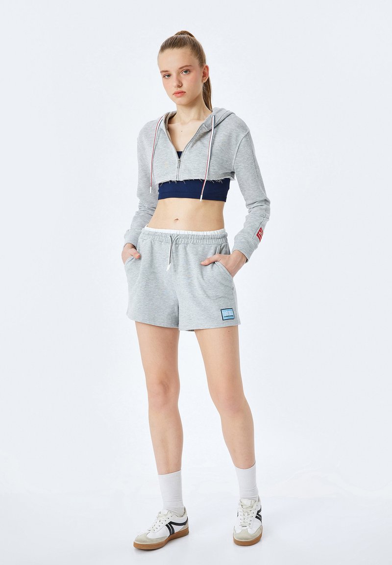 Młoda kobieta stojąca z rękami w kieszeniach, ubrana w szary, krótki hoodie, granatowy stanik sportowy, szare szorty, białe skarpetki i białe sneakersy.