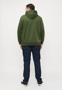 Green hoodie with a hood, ribbed cuffs, and hem. Worn with dark blue jeans and black footwear, viewed from the back against a white background.
