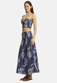 Modelo con un top corto sin mangas de flores y una falda larga a juego, de pie sobre un fondo liso, mostrando un look veraniego relajado.