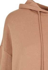 Hoodie od mekane, svijetlosmeđe tkanine. Ima teksturirani završetak, prostranu kapuljaču i vidljive vezice.